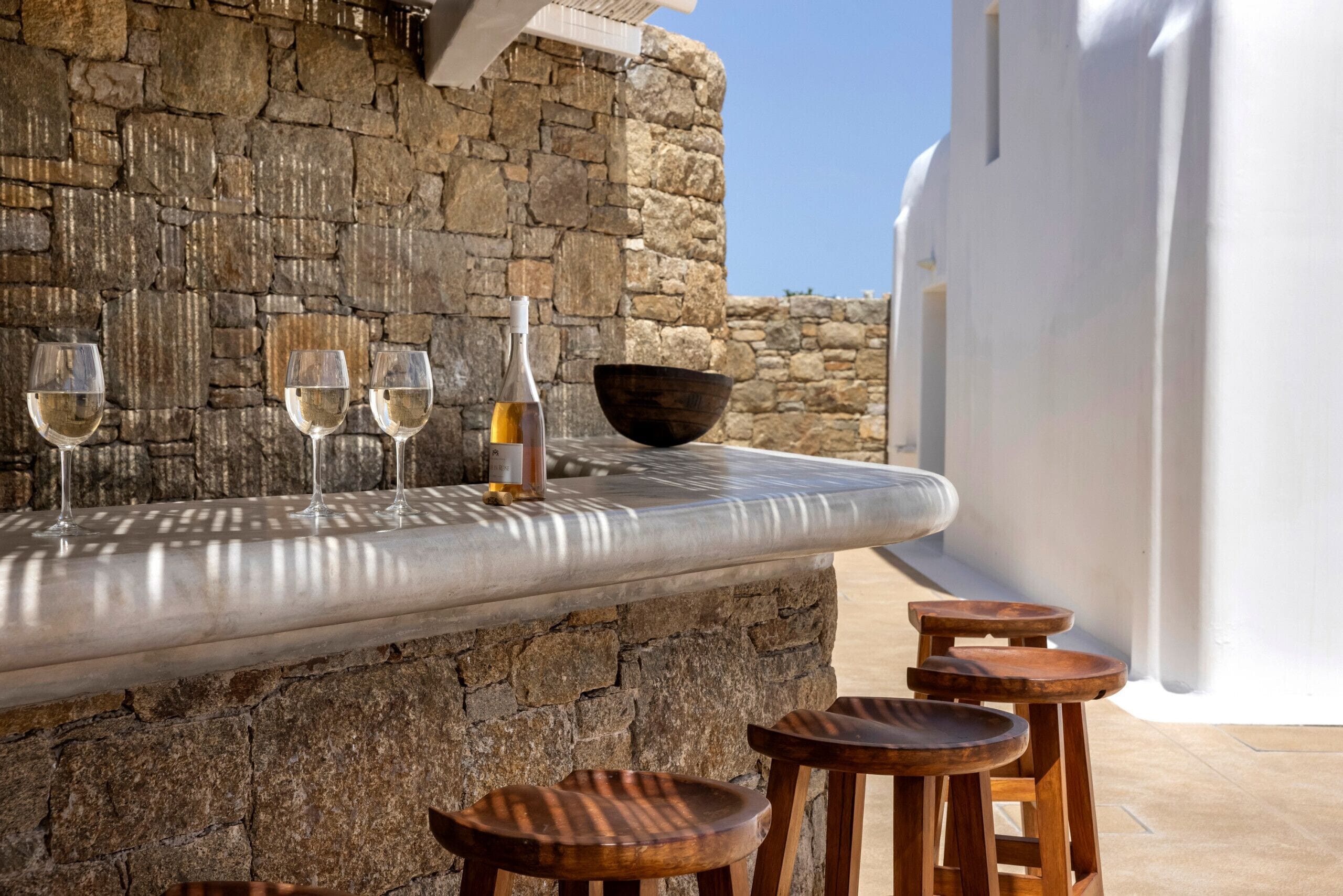 A stone wall bar area with three glasses of white wine, a bottle of wine, and a wooden bowl.