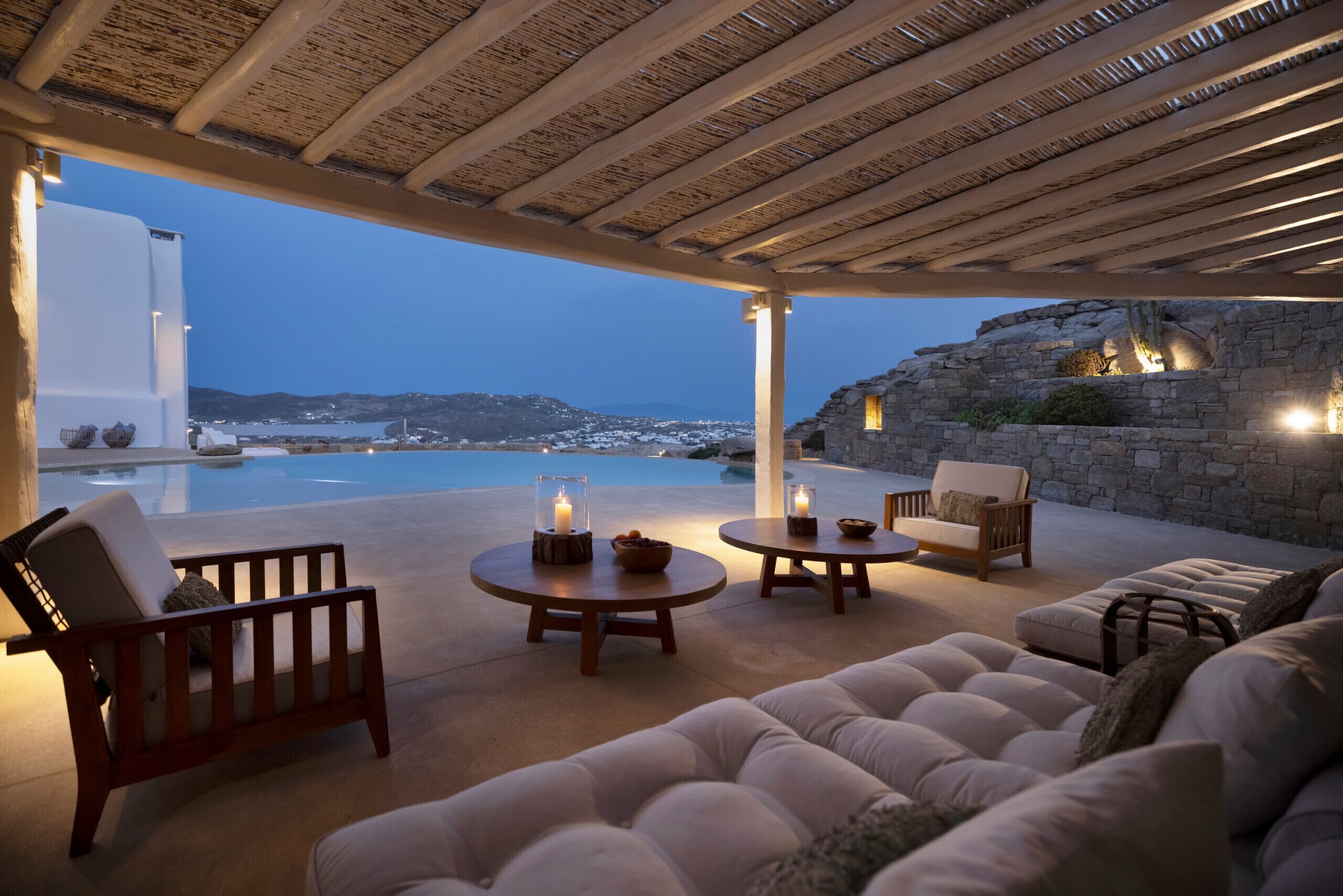 A cozy outdoor seating area with a view of the sea and mountains, featuring a wooden pergola, comfortable seating, and a stone wall.