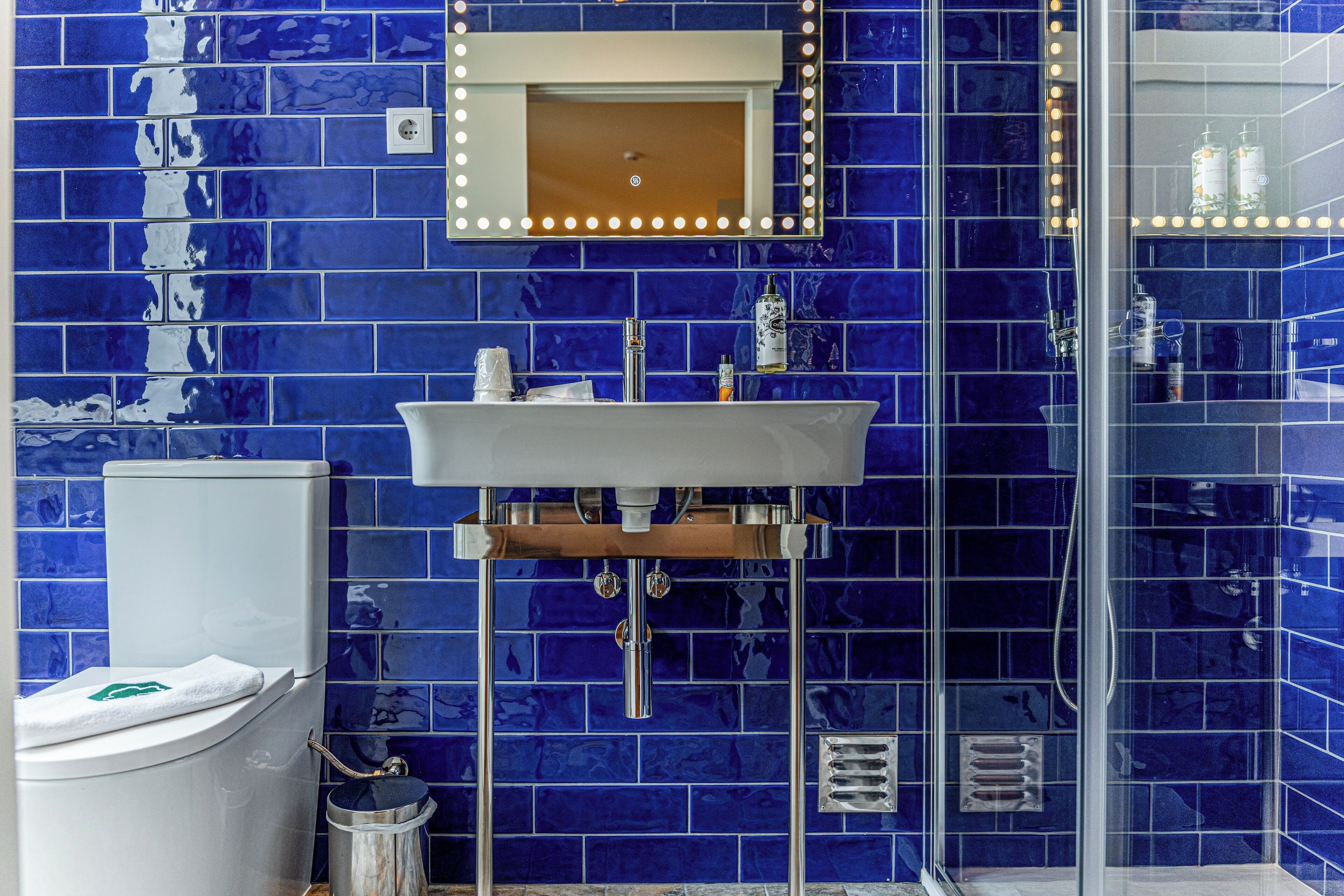 A bathroom with a blue-tiled wall, a white sink, a mirror with lights, and a glass shower enclosure.