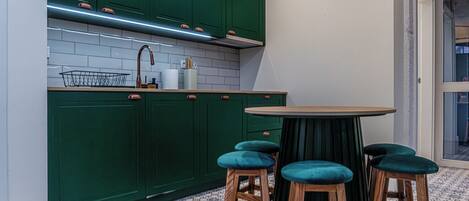 Una cocina con armarios verdes, un salpicadero de azulejos blancos, un grifo de cobre y una mesa redonda con cuatro taburetes.