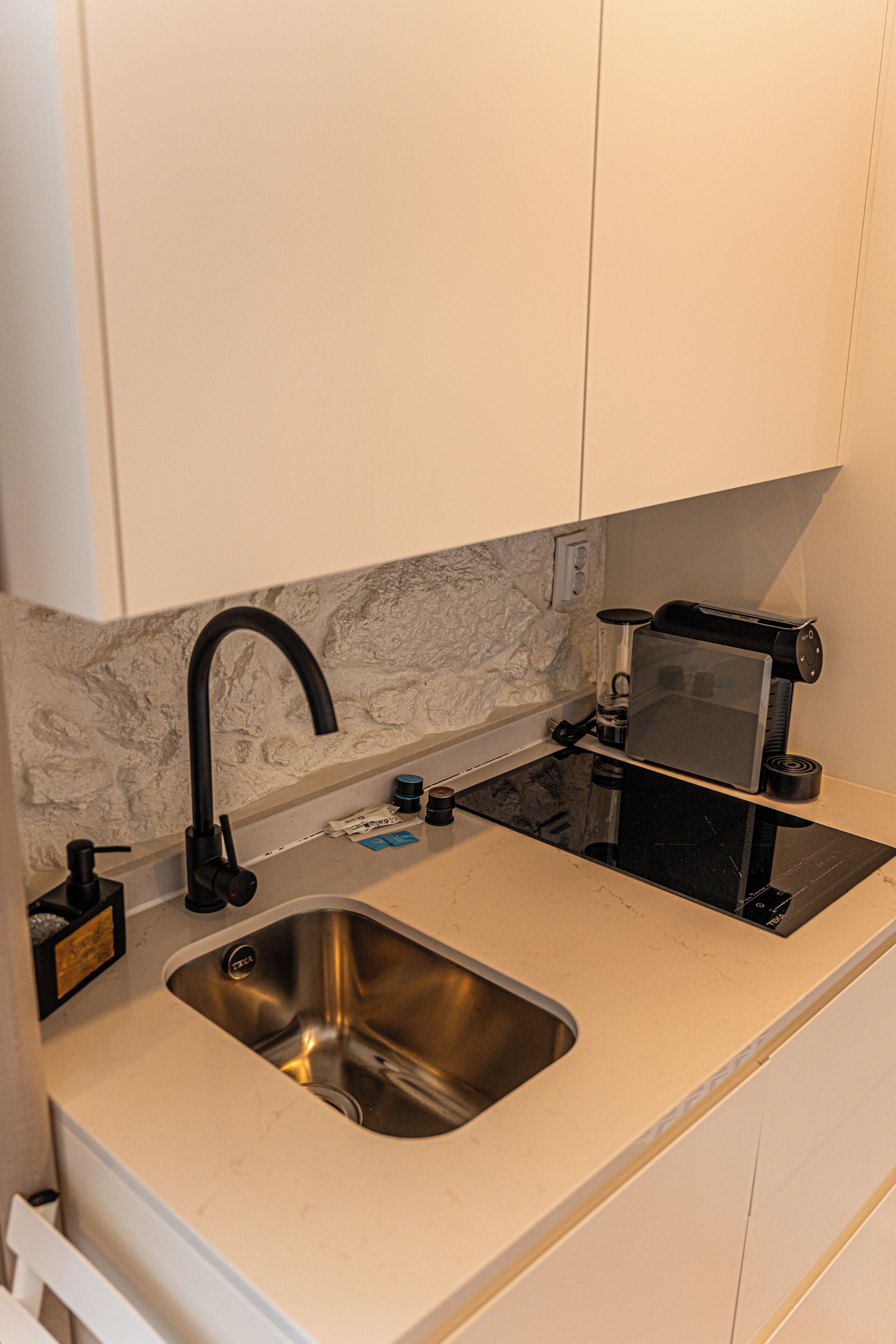 Cocina compacta con pileta de acero inoxidable, grifo negro, cafetera y protector de encimera de mármol.