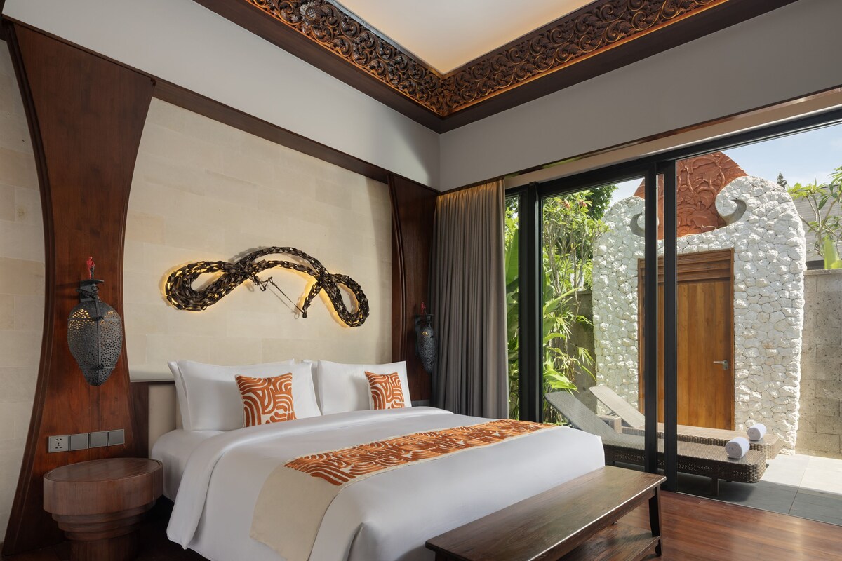 A spacious bedroom with a large bed, a bench, a wall-mounted light fixture, and a view of the outdoors.