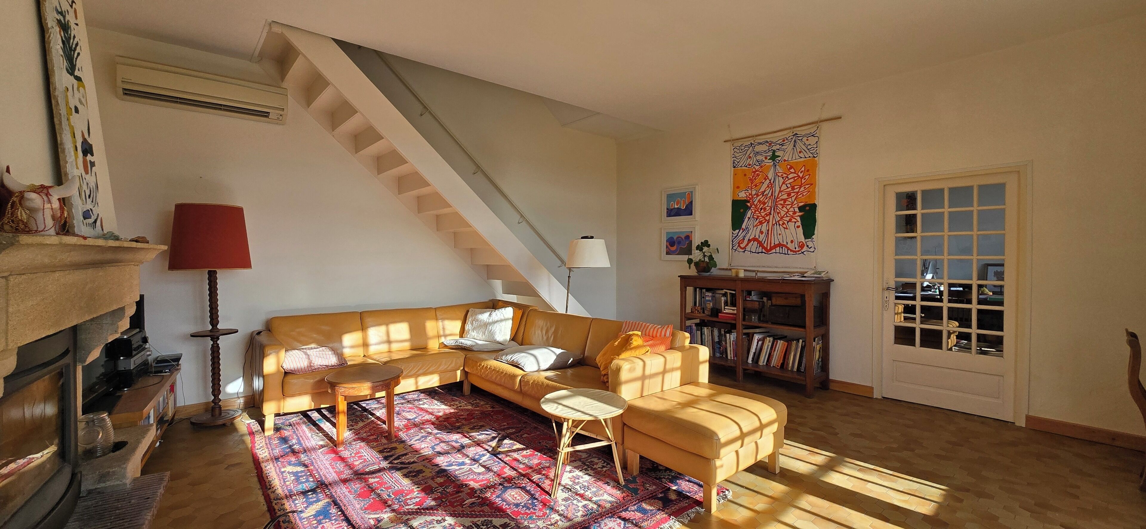 A living room with a wooden staircase, a sofa set, a patterned rug, a bookshelf, and a door with glass panels.