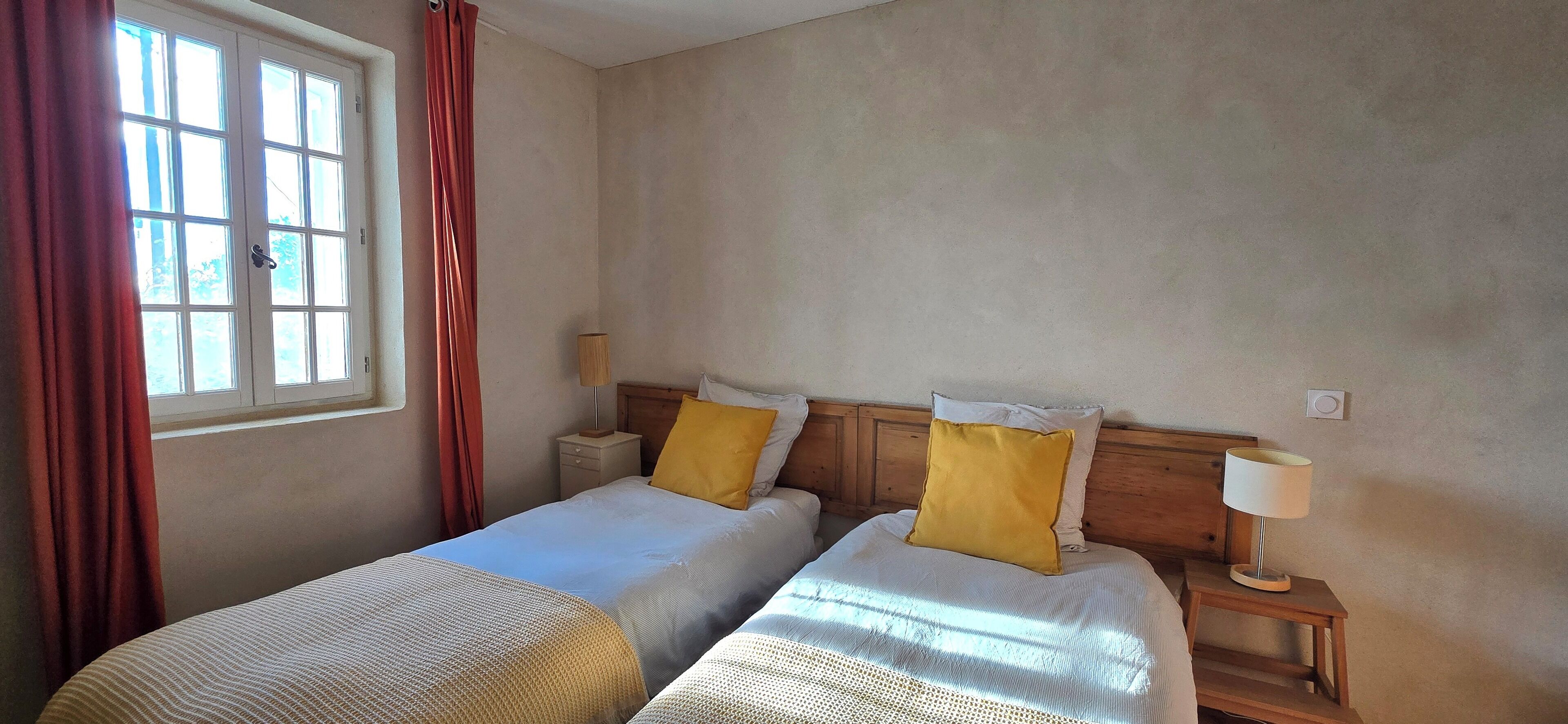 A hotel room with two beds, a window with curtains, a bedside table with a lamp, and a wall-mounted light switch.