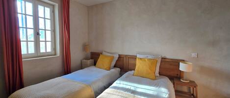 A hotel room with two beds, a window with curtains, a bedside table with a lamp, and a wall-mounted light switch.