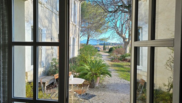 Une vue à travers une fenêtre donnant sur une cour avec un chemin de gravier, un banc en bois et une table entourée de chaises.