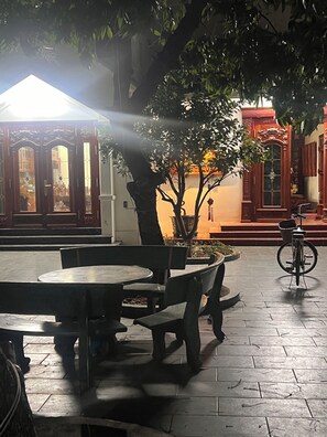 Un espacio al aire libre con mesas y bancos, rodeado de árboles y un edificio con puertas decorativas.