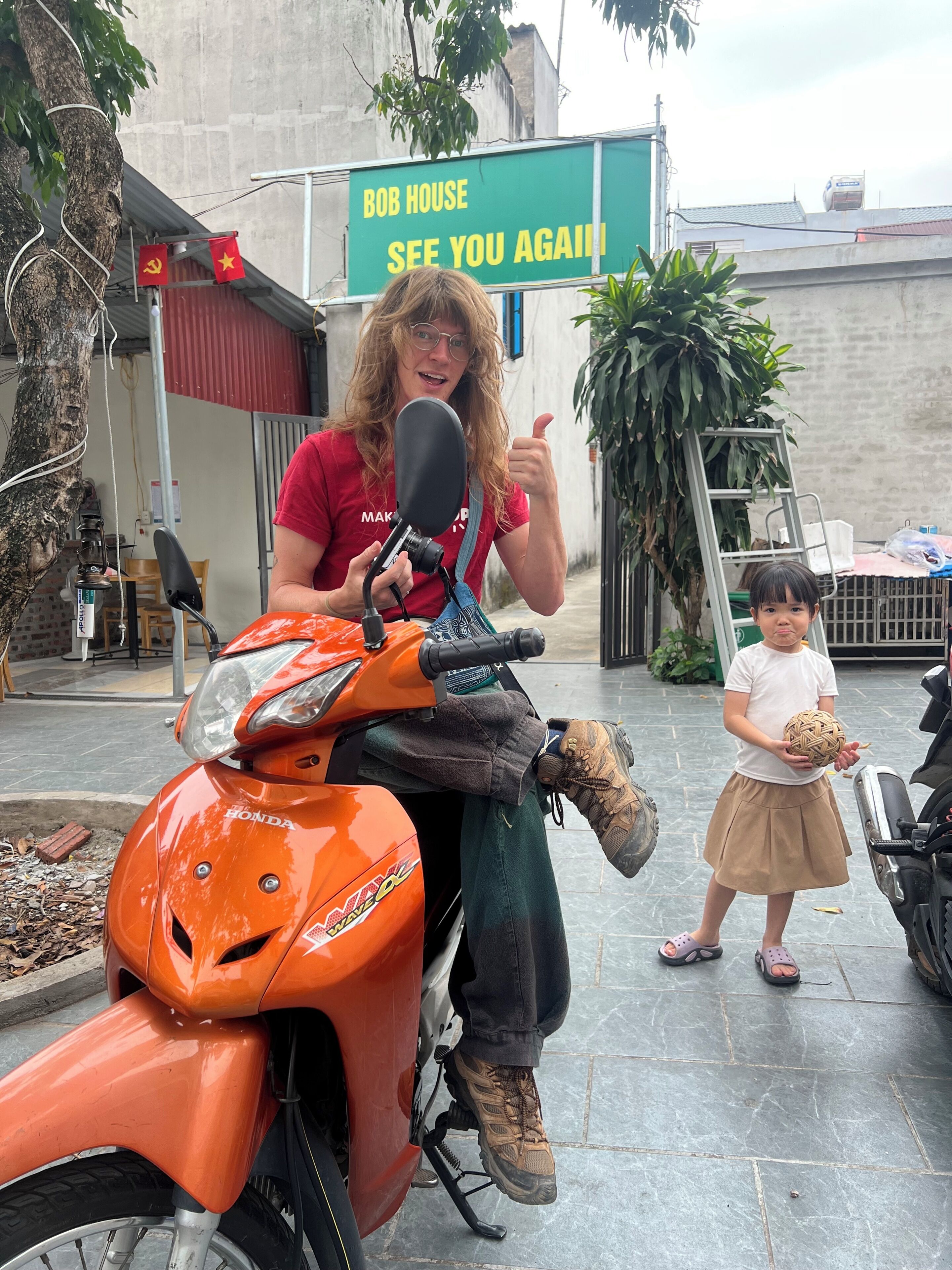 A person sitting on an orange motorcycle with a sign in the background that reads 'BOB HOUSE SEE YOU AGAIN'.