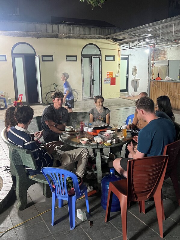 Un groupe de personnes est assis autour d’une table, devant divers plats, dans un cadre extérieur avec un bâtiment en arrière-plan.