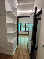 Um corredor com um guarda-roupa branco, soalho de madeira e uma porta de vidro que dá para uma varanda com vista para edifícios e árvores.