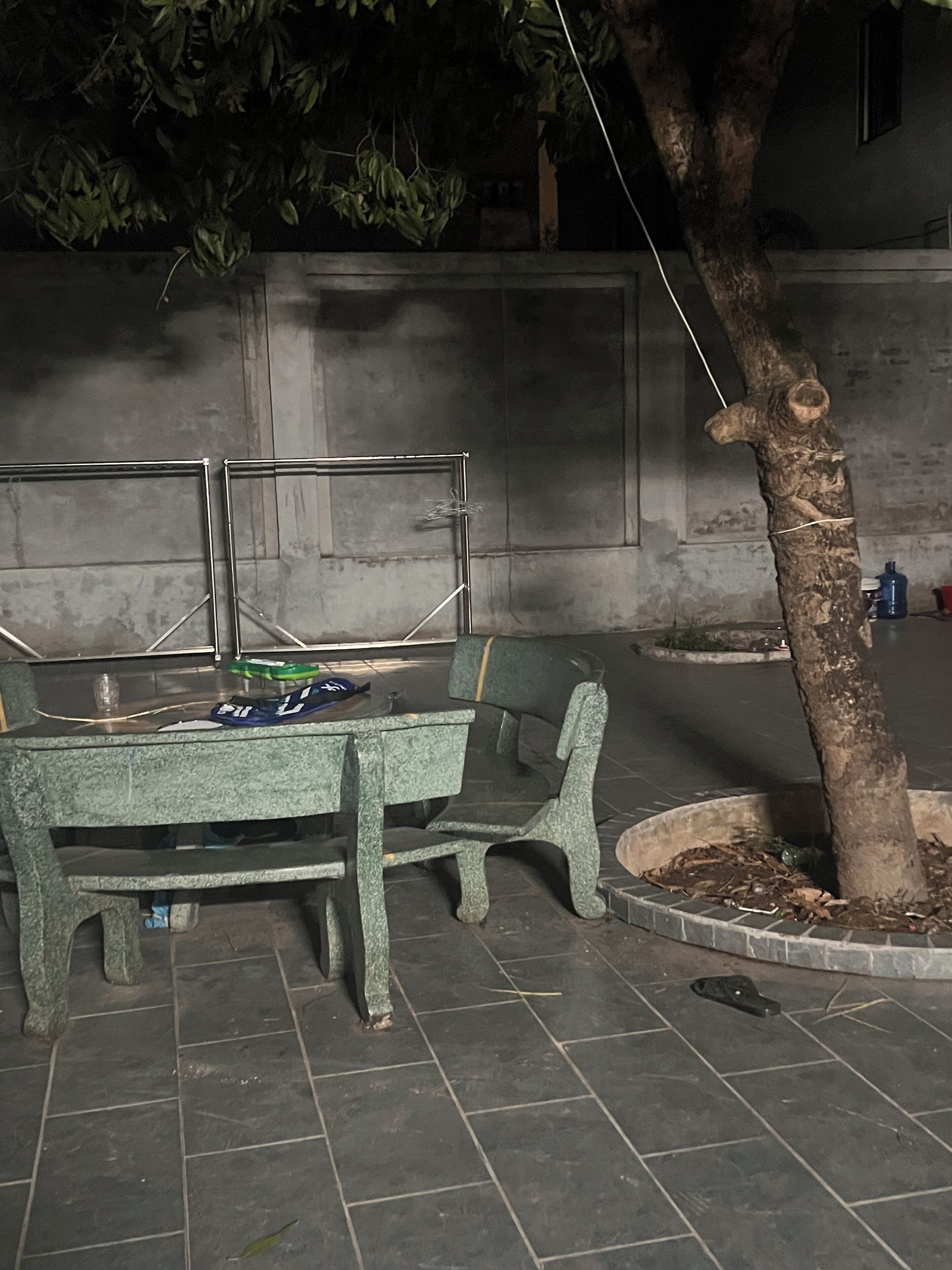 Un banco y una mesa en un espacio al aire libre, con un árbol y un muro de hormigón al fondo.