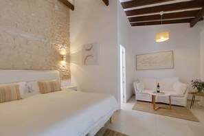 A bedroom with a stone wall, a bed, a white sofa, a small table with a bottle and glasses, and a painting on the wall.