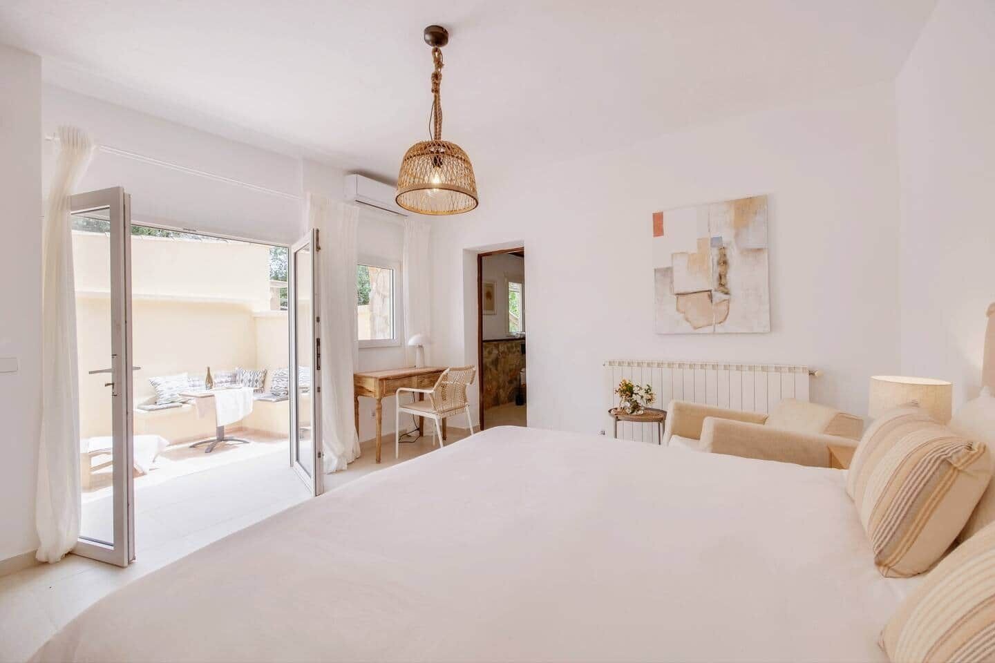 Ein geräumiges Schlafzimmer mit einem großen Bett, einem Schreibtisch und einem Stuhl, einem kleinen Tisch mit einer Lampe sowie Blick ins Freie.