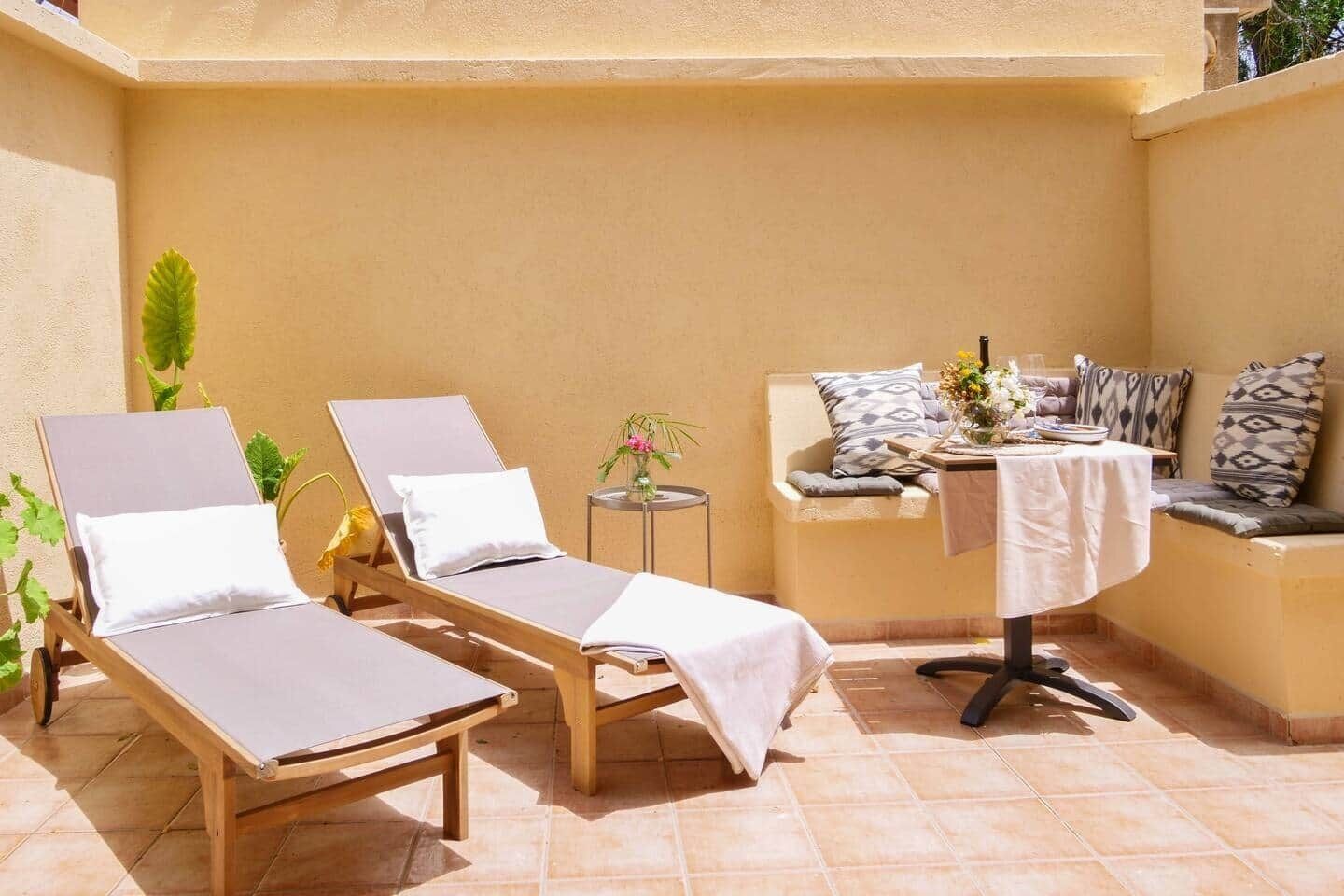 Un espace extérieur ensoleillé, aménagé avec deux fauteuils confortables, une petite table ronde et un banc rembourré agrémenté de coussins à motifs.
