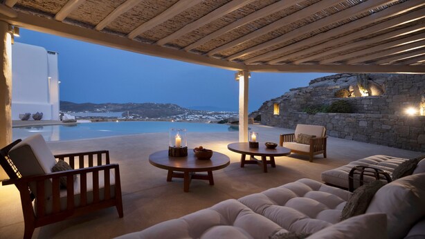 Ein gemütlicher Sitzbereich im Freien mit Blick auf das Meer und die Berge, mit einer Holzpergola, bequemen Sitzgelegenheiten und einer Steinmauer.
