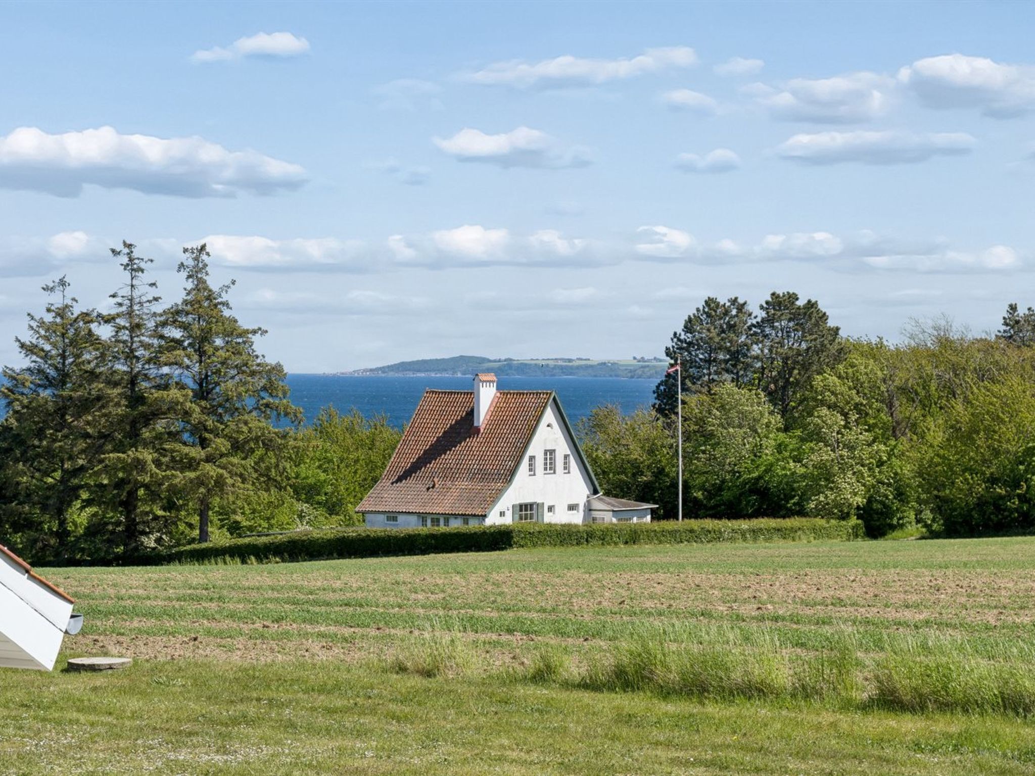 A house with a sloped roof, surrounded by a grassy field and trees, with a view of the sea in the background.