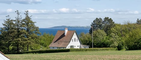 A house with a sloped roof, surrounded by a grassy field and trees, with a view of the sea in the background.