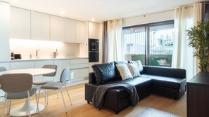 A modern living room with a black leather sofa, a dining table with chairs, and a kitchen area with white cabinets and a built-in oven. - Golden Comfort w/ Balcony by LovelyStay (Porto)