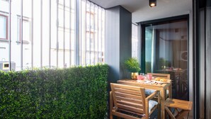 Ein Balkon mit Holztisch, gedecktem Frühstück, einer Topfpflanze und Blick auf die Stadt durch große Fenster.