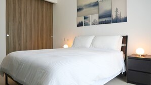 Ein ordentlich bezogenes Bett mit weißen Laken, ein Holzschrank und ein Nachttisch mit Lampe.