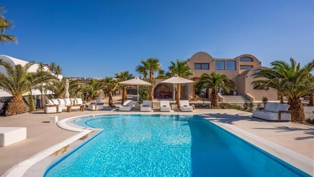 Ein Pool mit Liegestühlen und Sonnenschirmen, umgeben von Palmen und einem Gebäude mit einer geschwungenen Fassade.