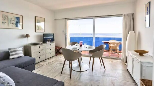 Een moderne woonkamer met een bank, een eettafel met stoelen, een televisie en een groot raam met uitzicht op zee.