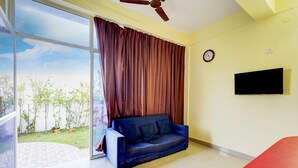 Ein Zimmer mit blauem Sofa, einem an der Wand befestigten Fernseher und einem großen Fenster mit Vorhängen.