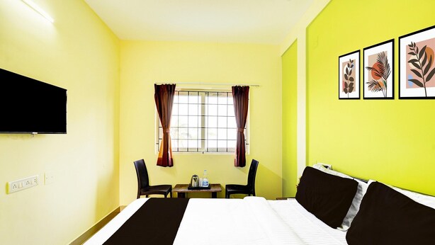 Ein Hotelzimmer mit Bett, einem an der Wand montierten Fernseher, einem Fenster mit Vorhängen, einem kleinen Tisch mit Wasserkocher und gerahmten Bildern an der Wand.