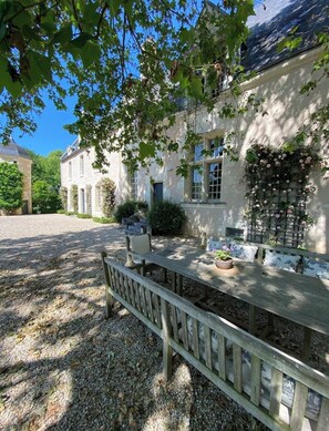 A courtyard with a wooden bench, a stone wall, and a building with a window and a door. - Suite d'hôtes - Domaine de Poulaines (Poulaines)
