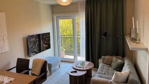 A living room with a sofa, armchair, and a table. There is a large window with curtains, a ceiling light, and artwork on the wall. - Modern apartment in the heart of Oslo with balcony (Oslo)