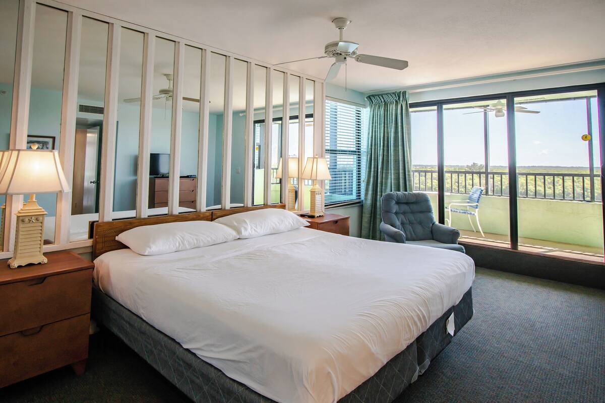 Een hotelkamer met een groot bed, een plafondventilator, uitzicht op de omgeving en een balkon met een stoel.