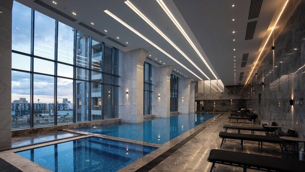 En moderne indendørs swimmingpool med store vinduer, et loungeområde med bænke og udsigt til bybilledet.