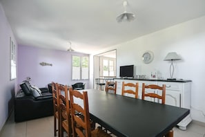 Un espace repas comprenant une longue table et des chaises en bois, un canapé, un téléviseur et un coin cuisine avec des placards et une horloge.