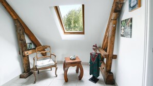 Ein Zimmer mit schrägen Wänden, Holzbalken, einem kleinen Tisch mit Kissen, einem Sessel und einer Statue.
