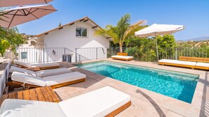 Un área de piscina con sillas de descanso, sombrillas y una casa al fondo.