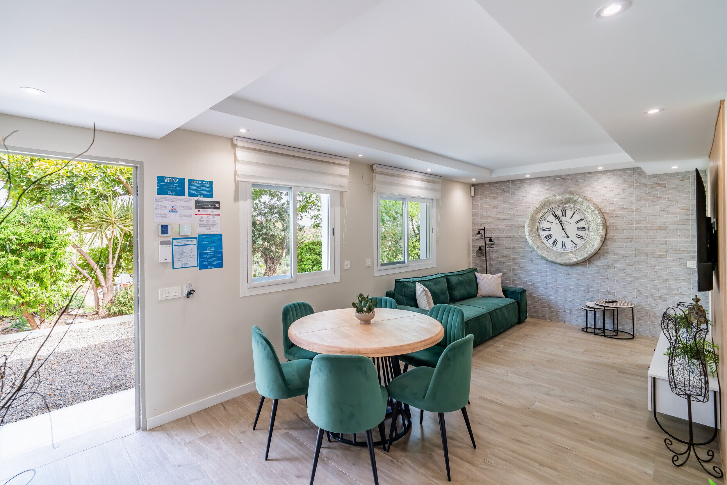 Un espace de vie moderne avec une table ronde en bois, des chaises recouvertes d’un tissu vert, un canapé vert et une grande horloge murale.