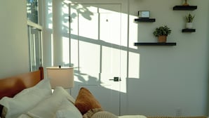Un dormitorio con cama, almohadas, lámpara de noche, ventana y puerta.