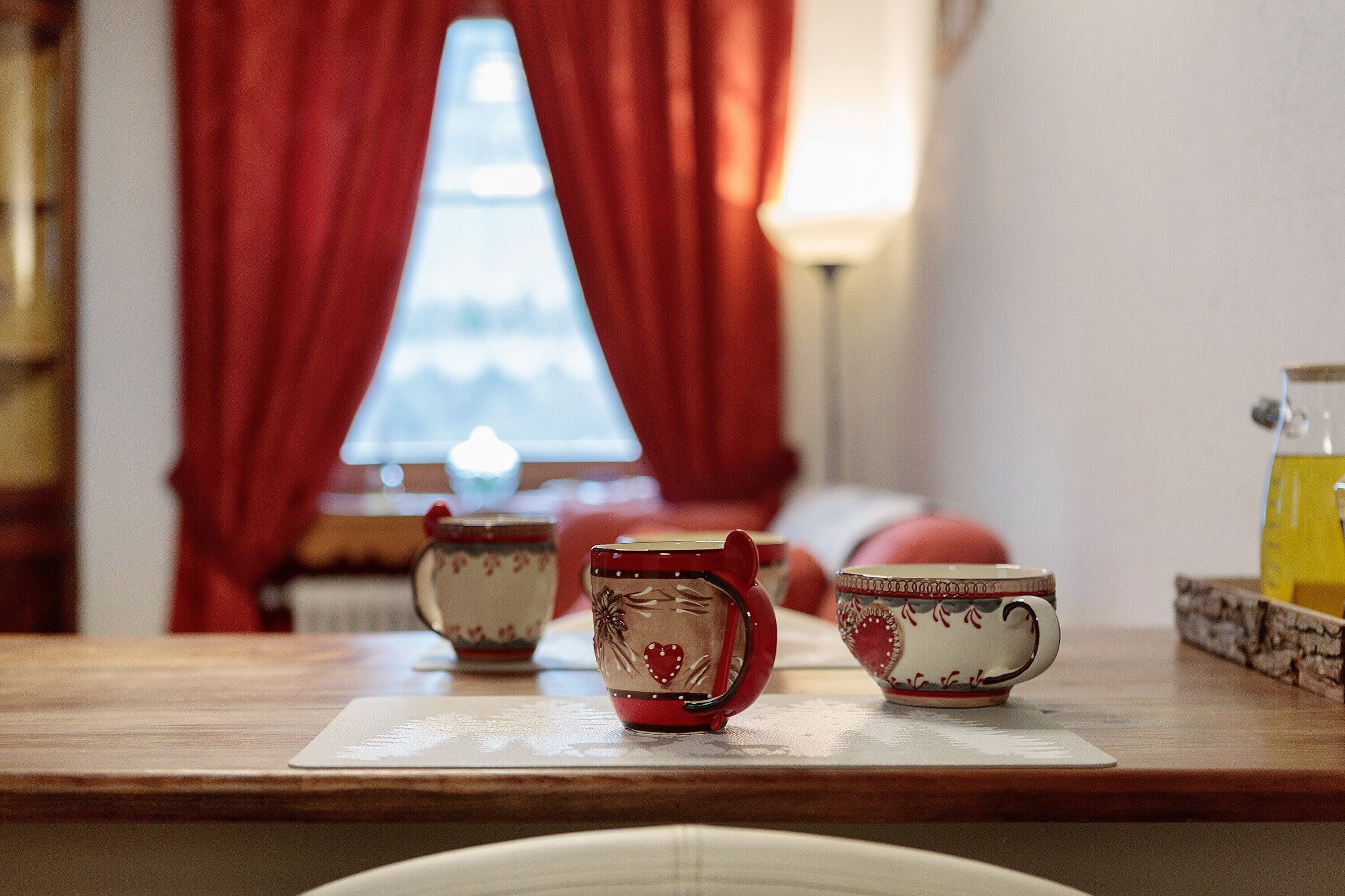 A table with three ceramic cups and a teapot, a window with red curtains, and a radiator in the background.