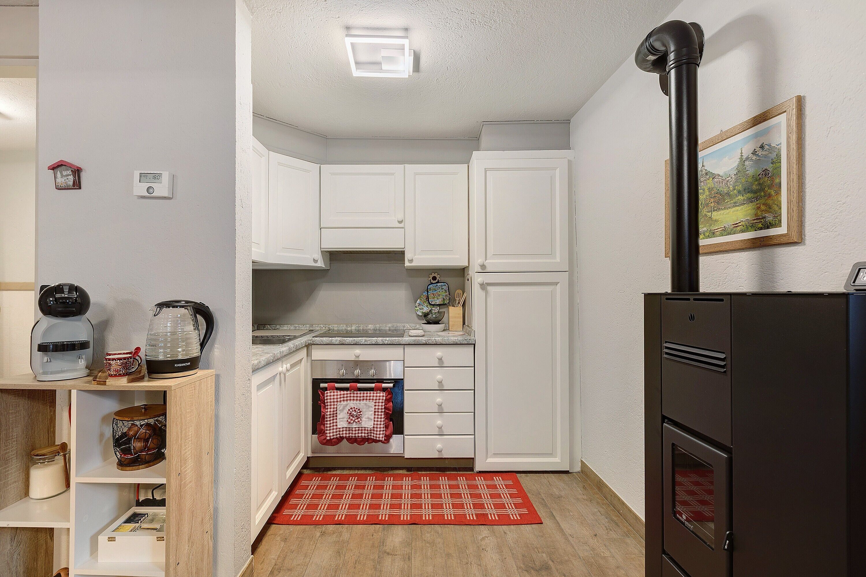 A compact kitchen with white cabinets, a black refrigerator, a coffee maker, a kettle, and a red patterned rug.