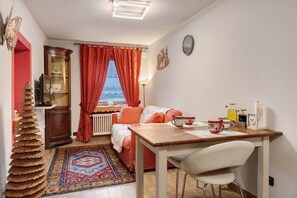 A cozy room with a wooden table, a sofa, a radiator, a window with red curtains, and a clock on the wall.