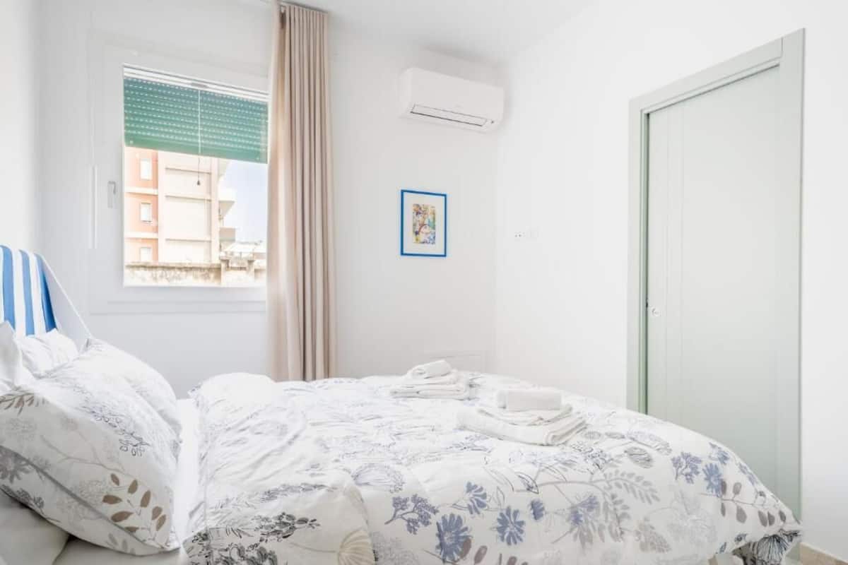 Quarto de hotel com cama, janela com cortinas, ar condicionado, porta e quadro na parede.