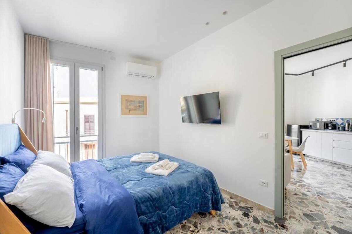Quarto com cama, TV fixada na parede, varanda com vista e área de cozinha visível através de uma porta aberta.