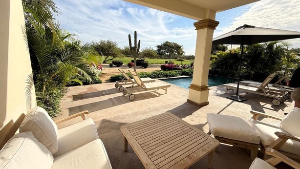 Un espacio de estar al aire libre con techo, sillones cómodos y una mesa, con vista a una piscina y jardín con cactus y palmeras.