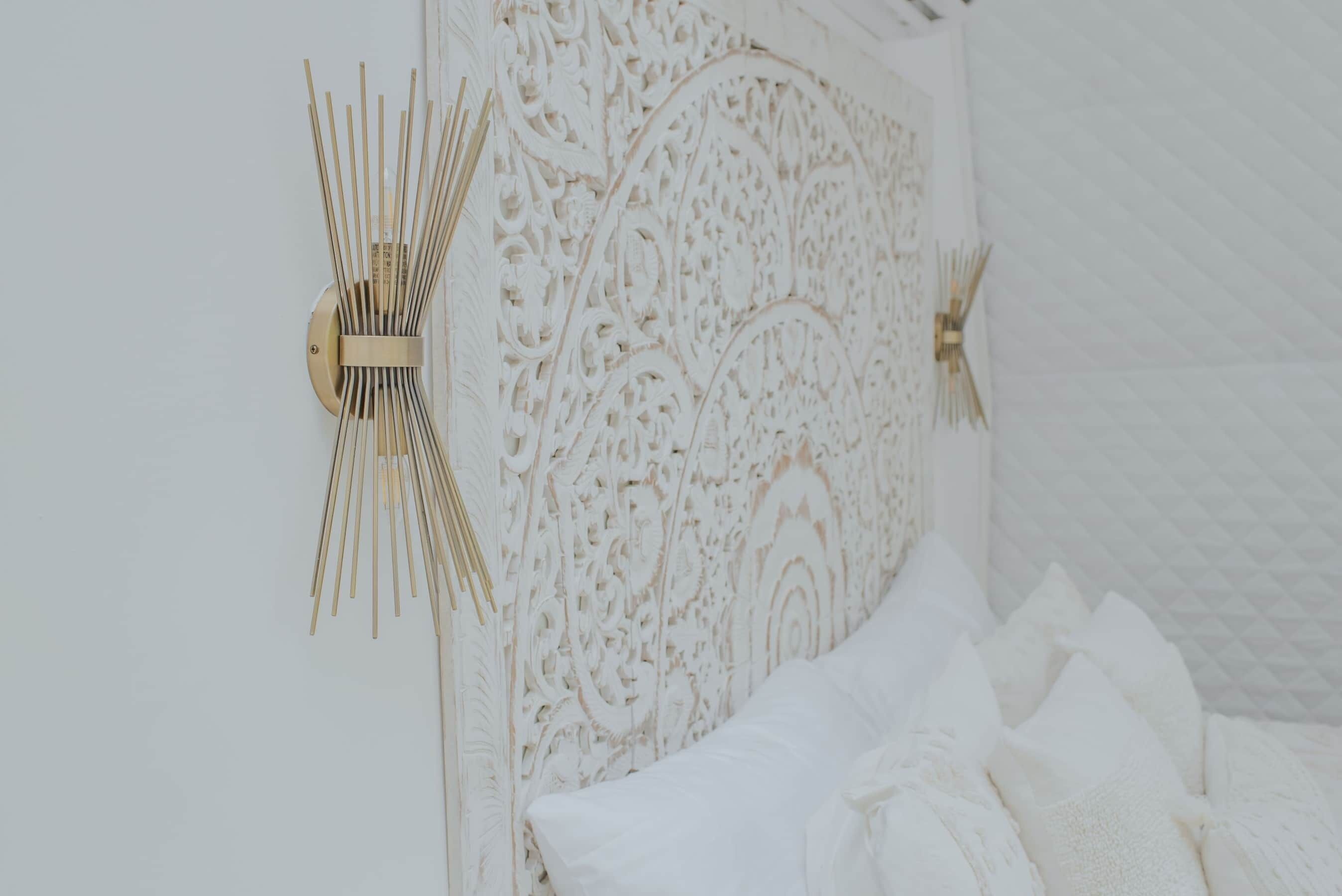A white, intricately carved headboard with a gold accent, a white bed with multiple pillows, and a white wall.