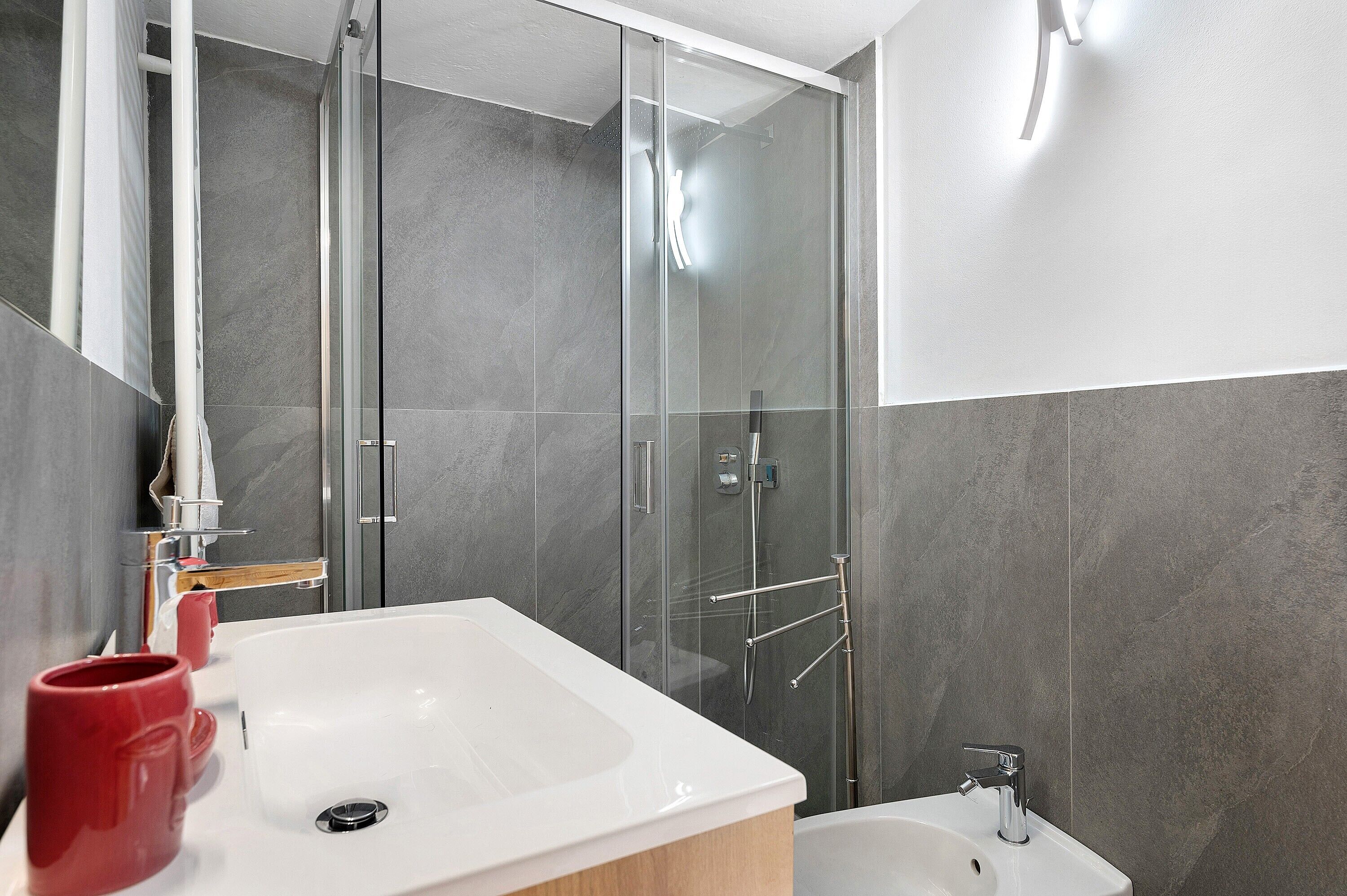 Un baño moderno con cabina de ducha de vidrio, un lavamanos blanco y una taza roja sobre el mostrador.