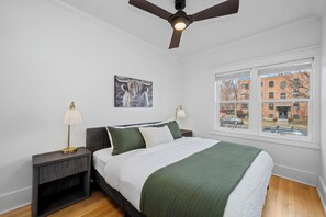 Ein Schlafzimmer mit Bett, Nachttischen, Deckenventilator, einem Fenster mit Blick auf Gebäude und einem gerahmten Bild an der Wand.