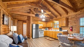 Interior de una cabaña de troncos con techo de madera, un comedor con mesa y sillas, una cocina con refrigerador y estufa, y una zona de estar acogedora con sofá y cojines.