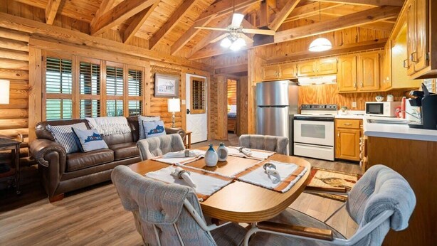 Un acogedor interior de cabaña de troncos con una mesa de comedor de madera, un sofá y una zona de cocina.