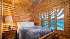 Dormitorio de cabaña de madera con cama, mesita de noche, lámpara y ventana grande con contraventanas.
