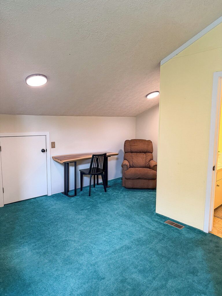 Una habitación con alfombra color turquesa, un escritorio de madera, una silla negra y un sillón reclinable marrón.